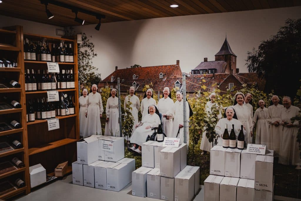 Wijnflessen op schappen en in dozen in een winkel; op een grote foto aan de muur is een groep vrouwen in witte gewaden te zien die in een wijngaard bij Breda staan, met historische gebouwen op de achtergrond – een knipoog naar nonnenwijn en het goede leven in het bos.