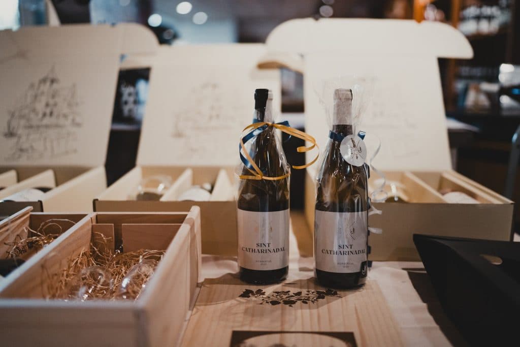 Twee flessen Sint Catharinadal nonnenwijn, versierd met strikken, staan op een tafel, omringd door geopende geschenkdozen en verpakkingsmateriaal – perfect om ‘het goede leven’ te vieren.