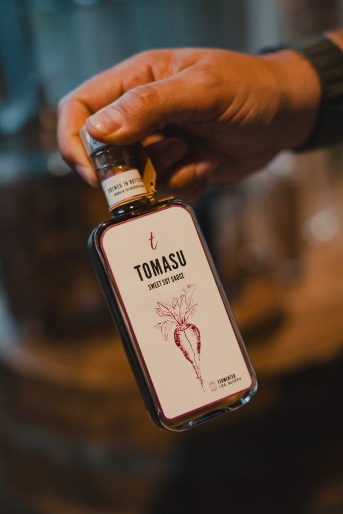 Een hand houdt een rechthoekige fles Tomasu Sweet Soy Sauce vast met een rood etiket waarop een radijsje is afgebeeld, waarmee de essentie van het goede leven in Breda perfect wordt weergegeven.