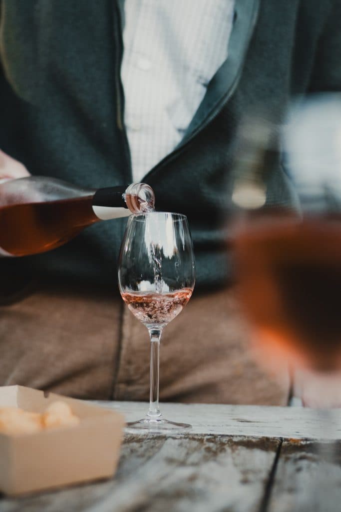 Iemand schenkt nonnenwijn rosé uit een fles in een wijnglas op een houten tafel, met hapjes op de voorgrond, waarmee het goede leven tijdens een ‘Bruisend Breda’-bijeenkomst wordt vastgelegd.