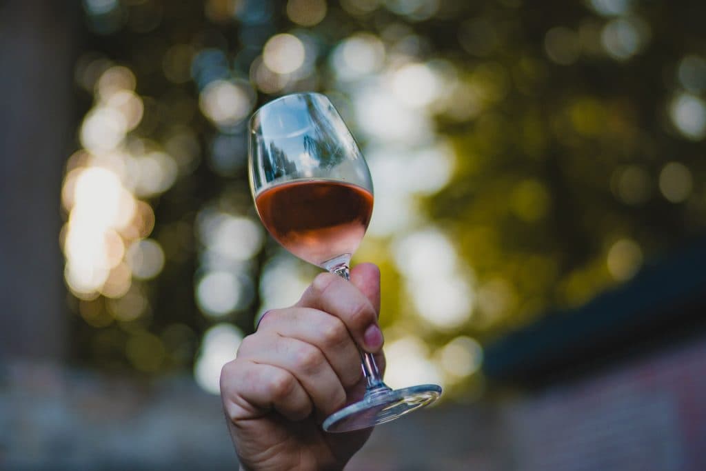 Een hand houdt buiten een glas Nonnenwijn rosé omhoog, tegen een wazige achtergrond van groen en geel gebladerte – een uitnodiging om te proosten op het goede leven.