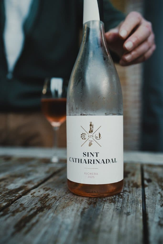 Iemand houdt een fles Sint Catharinadal Ricwera 2023 rosé – ook wel bekend als nonnenwijn – vast op een houten tafel, met op de achtergrond een wijnglas, waarmee de essentie van het goede leven wordt weergegeven.