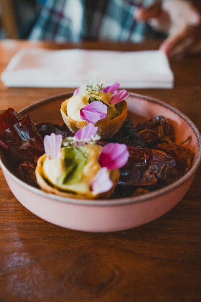 Een roze schaal met daarin twee kleine hapjes, gegarneerd met room en bloemblaadjes, omringd door gedroogde rode chilipepers, op een houten tafel – een prachtige presentatie die ‘het goede leven’ en de levendige smaken van Breda perfect weergeeft.