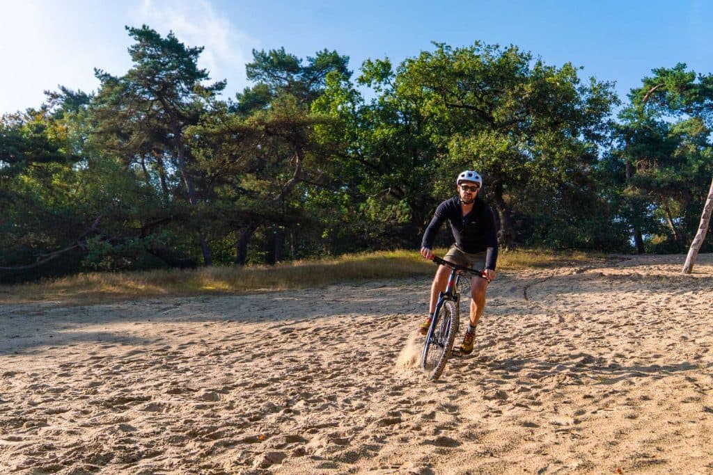 Iemand met een helm op fietst over zanderig terrein, met bomen en gras op de achtergrond en onder een gedeeltelijk bewolkte hemel, en geniet van het goede leven in de buurt van Bruisend Breda.