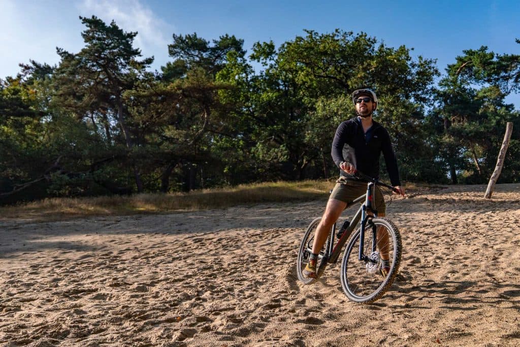 Iemand met een helm op rijdt op een mountainbike over een zandpad in de buurt van Breda, met bomen en een blauwe lucht op de achtergrond, en geniet met volle teugen van het goede leven.