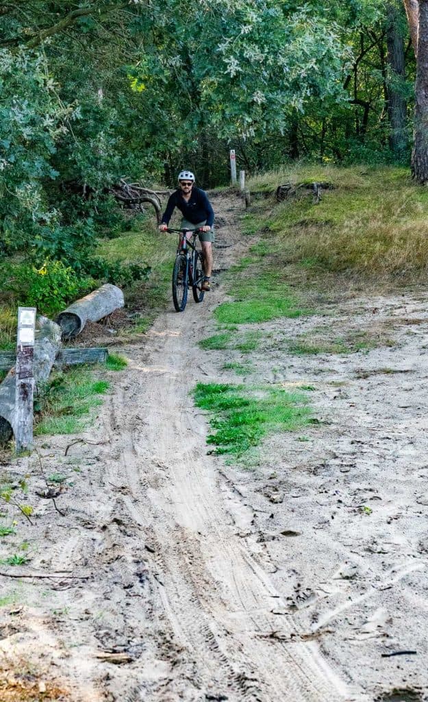Iemand met een helm op fietst over een onverhard pad door een bosrijke omgeving in de buurt van Breda, met aan weerszijden bomen en gras, en geniet van het goede leven te midden van de rust van de natuur.