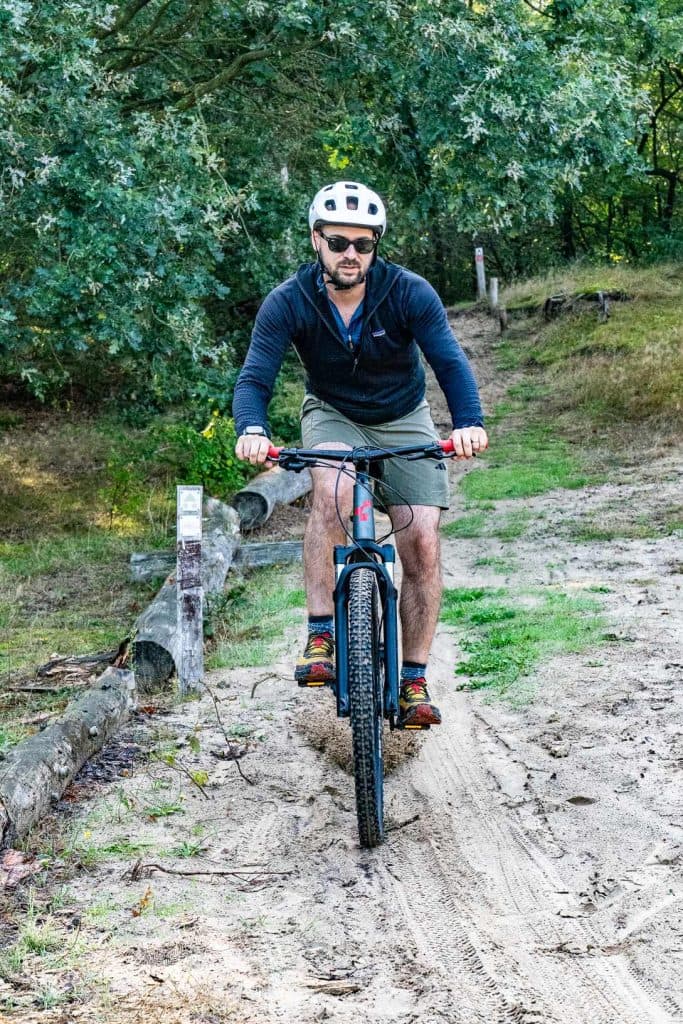 Een man met een helm en een zonnebril rijdt op een mountainbike over een zanderig pad, omringd door bomen en gras, en geniet van het goede leven midden in de natuur.