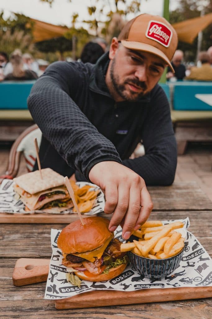 Een man die op een houten tafel naar frietjes grijpt naast een hamburger, met nog een broodje en mensen op de wazige achtergrond — een beeld van het goede leven in Breda, dat je misschien het best kunt genieten met een glas nonnenwijn.