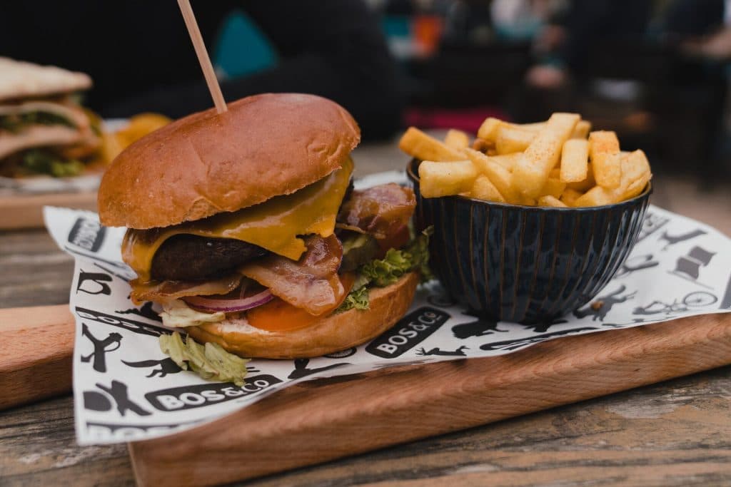 Een cheeseburger met sla, tomaat, spek en rode ui wordt geserveerd op een houten plank, samen met een schaaltje frietjes – perfect om in Breda van het goede leven te genieten, vooral in combinatie met een glas nonnenwijn.