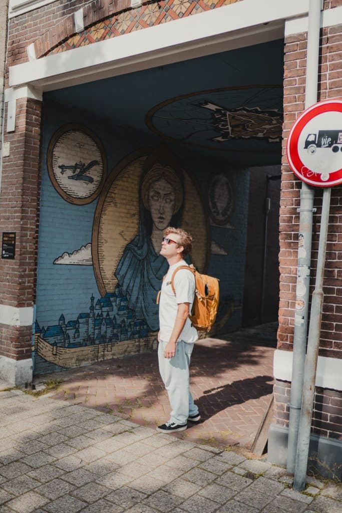 Een persoon met een bruine rugzak staat onder een boog in Breda en kijkt omhoog naar een muurschildering met daarop een vrouw, gebouwen en een vliegtuig – een levendige weergave van het goede leven. Rechts is een verkeersbord te zien.