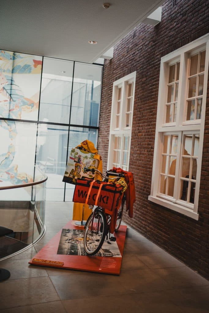 Binnen staat een fiets met een rode bezorgkist waarop ‘WOAH!’ staat, naast een etalagepop in een gele regenjas, tegen een achtergrond van bakstenen en glazen muren – een eigenzinnig tafereel dat het goede leven in het hart van Bruisend Breda perfect weergeeft.
