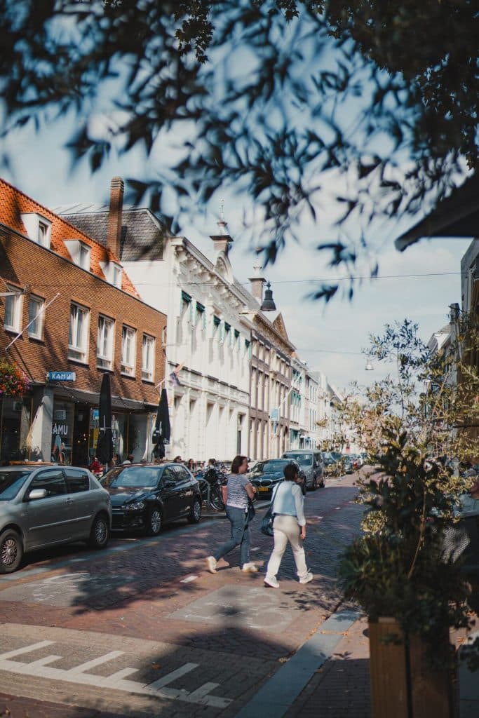 Op een zonnige dag steken twee mensen in Breda een met bakstenen geplaveide straat over, die wordt omzoomd door geparkeerde auto’s en winkels. Lommerrijke takken omlijsten het tafereel gedeeltelijk van bovenaf en geven een glimp weer van het goede leven.