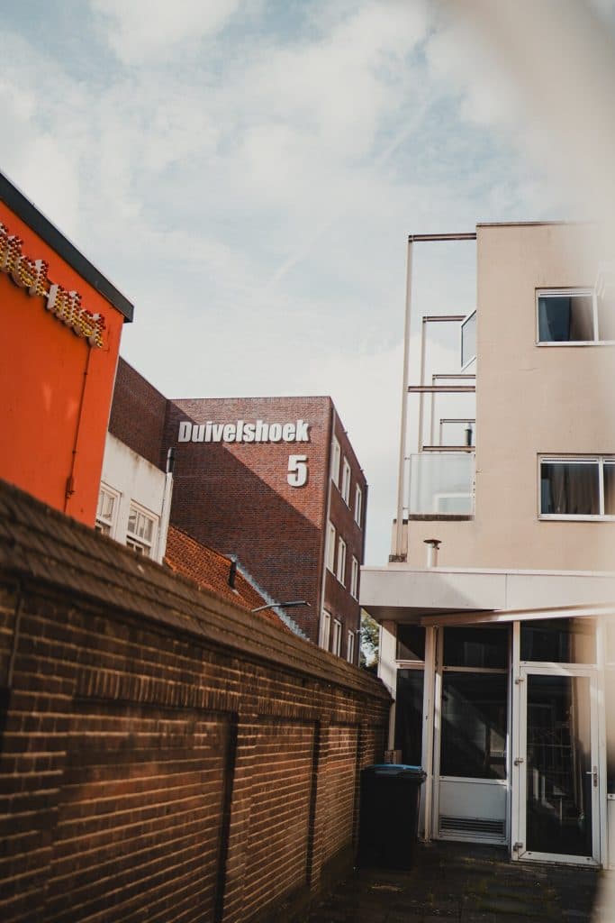 Achter een wit, modern gebouw en een bakstenen muur, onder een gedeeltelijk bewolkte hemel, is een bakstenen gebouw te zien met het opschrift ‘Duivelshoek 5’, dat de essentie van het goede leven in Breda weergeeft.