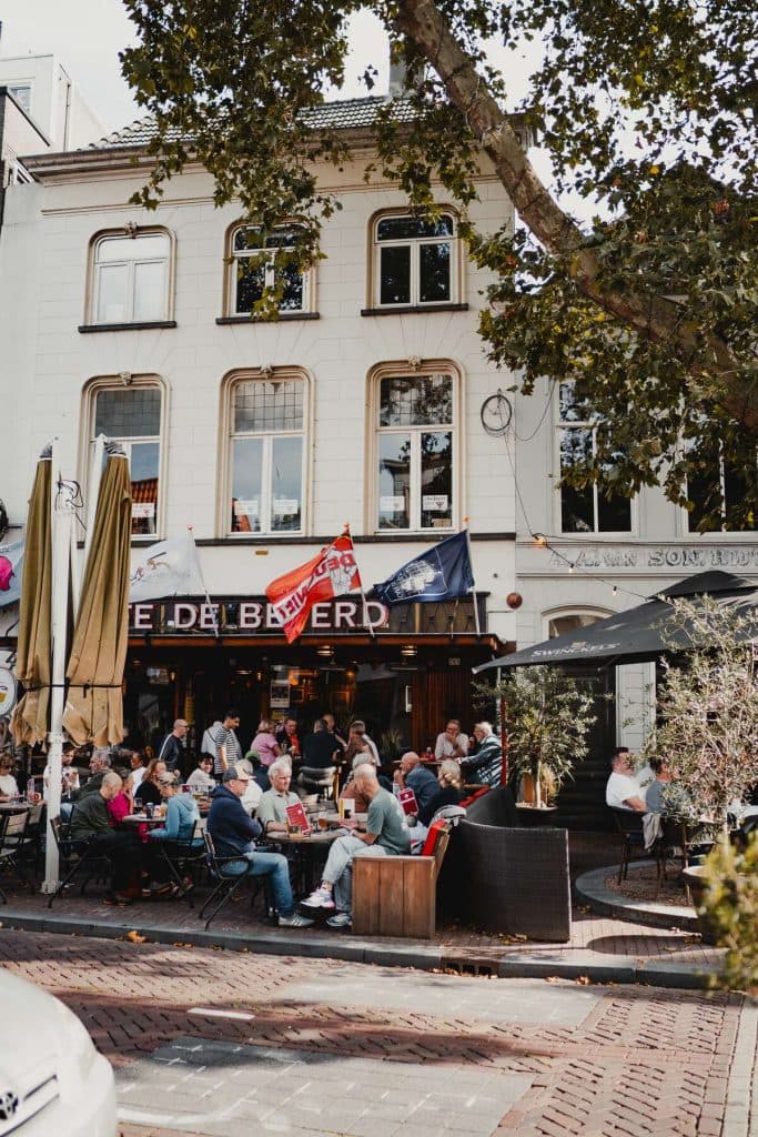 Mensen zitten aan tafeltjes buiten voor een druk café in een straat met kasseien en genieten met volle teugen van het goede leven, met bomen en een wit gebouw op de achtergrond. Boven de ingang van het café hangen vlaggen, wat bijdraagt aan de bruisende sfeer van Breda.