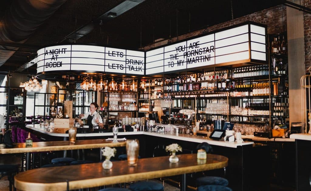 Modern barinterieur in Breda met planken vol flessen, hanglampen en grote borden boven de bar met speelse teksten over het goede leven. Achter de toog staat een barman, klaar om een glas nonnenwijn in te schenken, terwijl op de voorgrond tafels met bloemen uitnodigend zijn opgesteld.