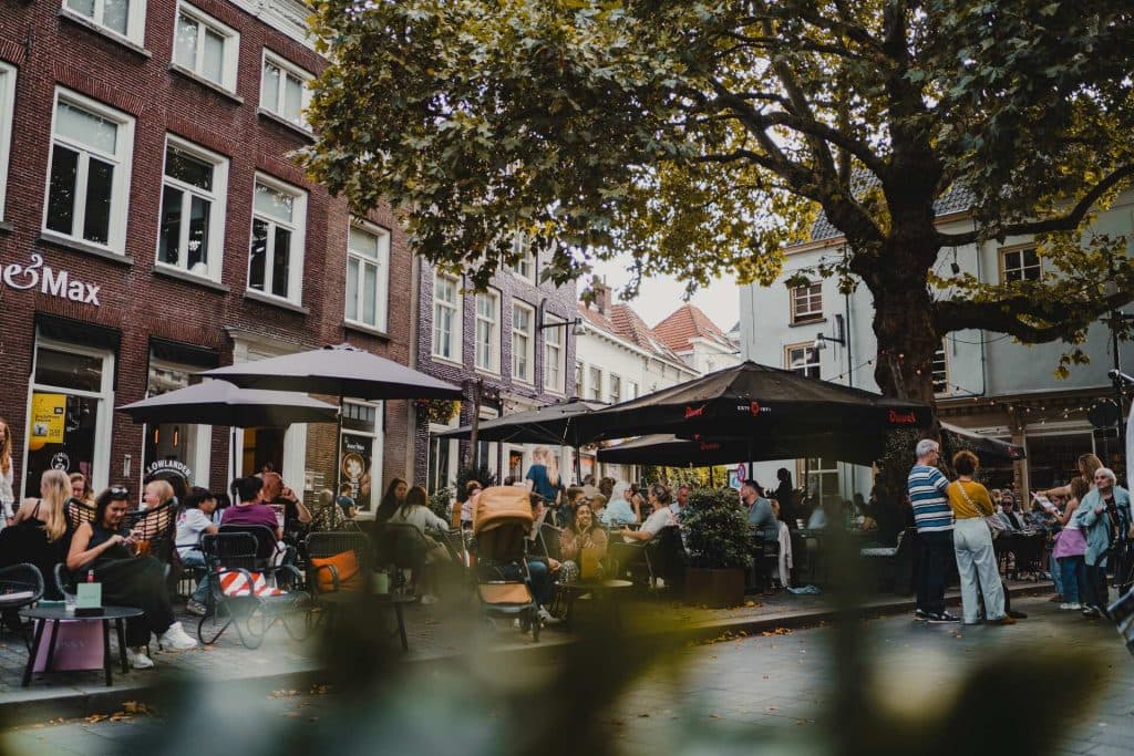 Overdag zitten mensen aan terrastafels onder parasols in een straat vol bakstenen gebouwen en grote bomen, waar ze genieten van het goede leven dat Bruisend Breda te bieden heeft.