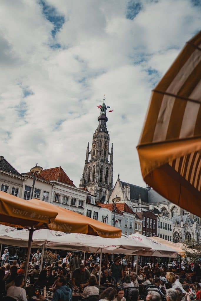 Een terras met drukbezette tafels onder parasols, gelegen in het hart van Breda, waar de sfeer van het ‘goede leven’ voelbaar is in elk gesprek en elke slok nonnenwijn, terwijl op de achtergrond een hoge, sierlijke kerktoren zich gracieus aftekent tegen een gedeeltelijk bewolkte hemel.