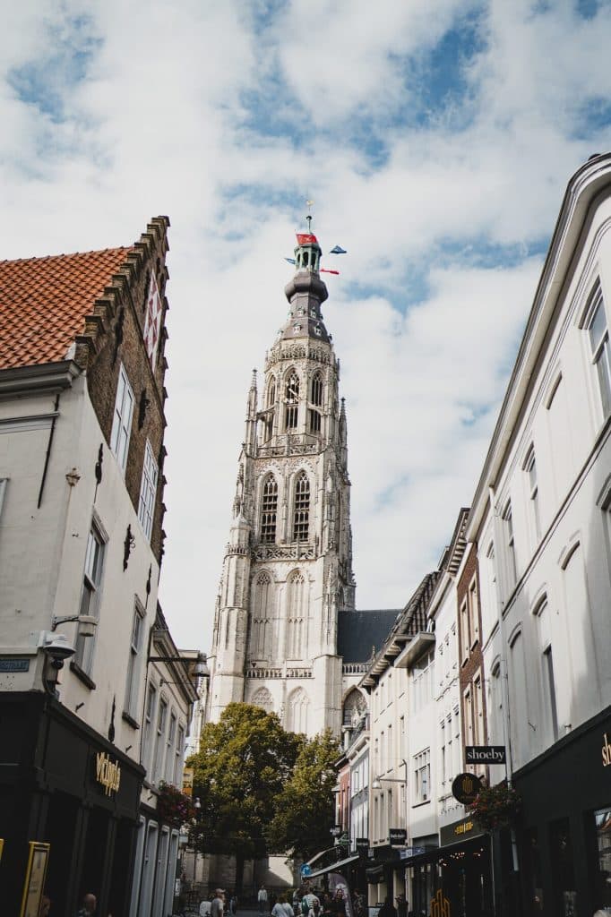 Een hoge gotische kerktoren torent uit boven de omliggende winkels en gebouwen in een straat in de stad onder een bewolkte hemel, en weerspiegelt daarmee de sfeer van ‘het goede leven’ waar Bruisend Breda om bekendstaat.