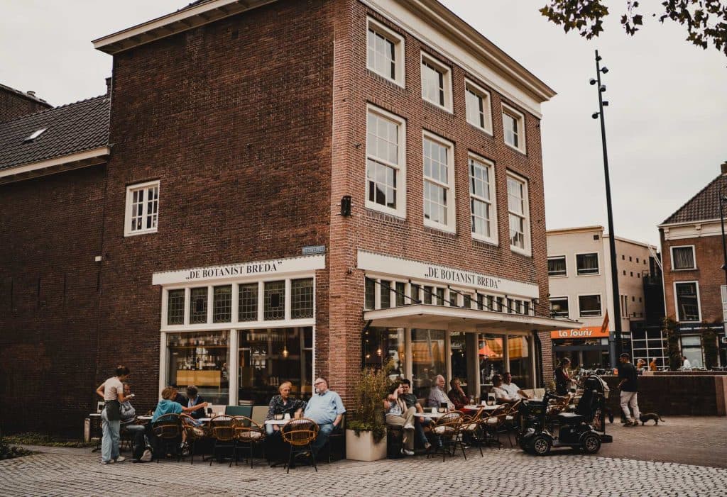 Mensen zitten aan de terrastafels van een hoekcafé genaamd ‘De Botanist Breda’ in een bakstenen pand aan een straat met kasseien, waar ze genieten van de bruisende sfeer in Breda terwijl ze nippen aan lokale nonnenwijn en het leven aan zich voorbij zien gaan.