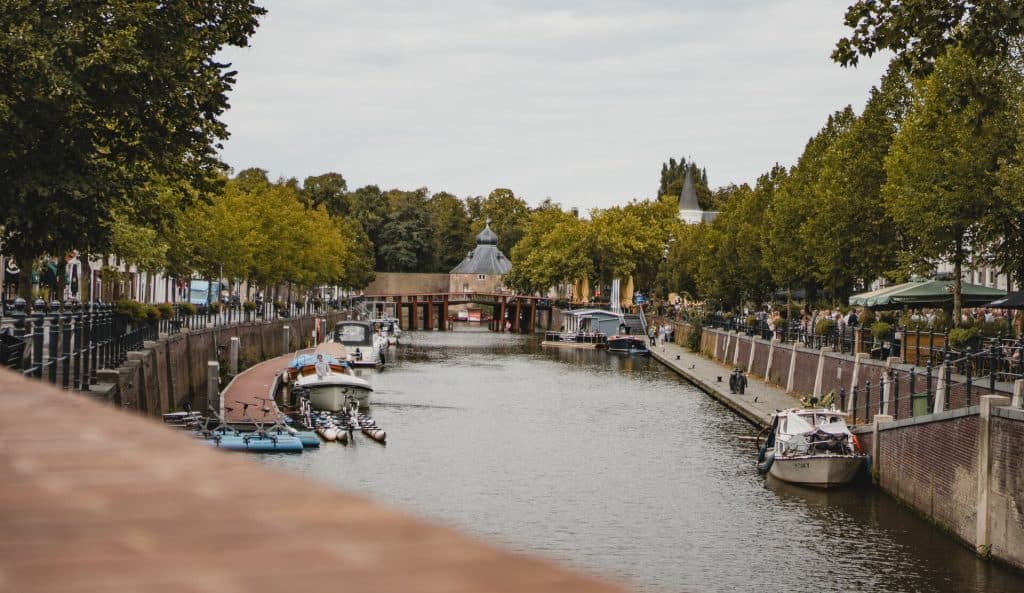 Een met bomen en boten omzoomd kanaal strekt zich uit richting een gebouw in de verte, terwijl voetgangers over de paden aan weerszijden lopen – een beeld dat de essentie weergeeft van ‘het goede leven’ waar Bruisend Breda om bekend staat.