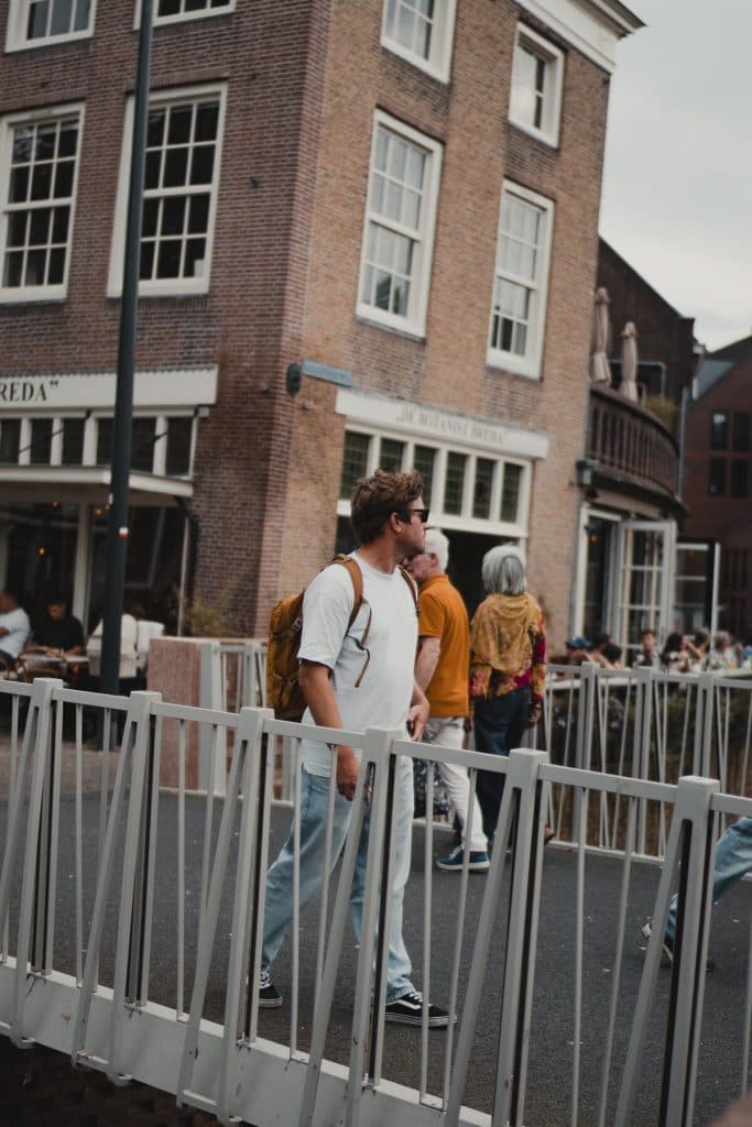 Een man met een zonnebril en een rugzak loopt over een witte brug in het stadscentrum van Bruisend Breda, met bakstenen gebouwen en mensen op de achtergrond, wat het goede leven perfect weergeeft.
