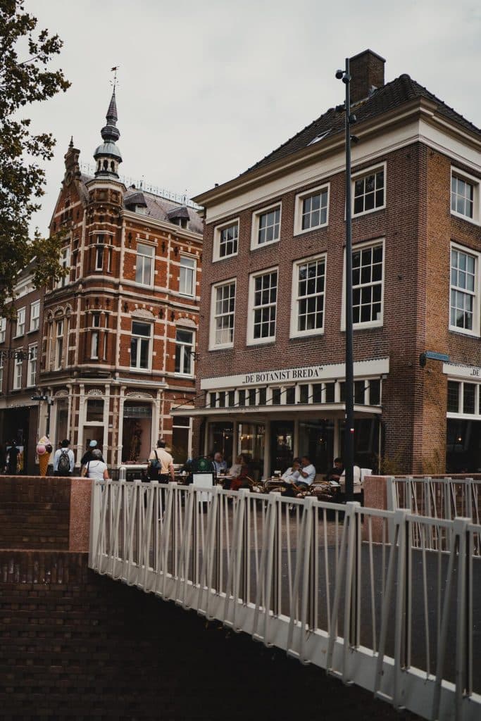 Mensen zitten aan terrastafels voor een café met de naam ‘The Botanist Breda’ en genieten van het goede leven te midden van historische bakstenen gebouwen en een witte reling op de voorgrond.