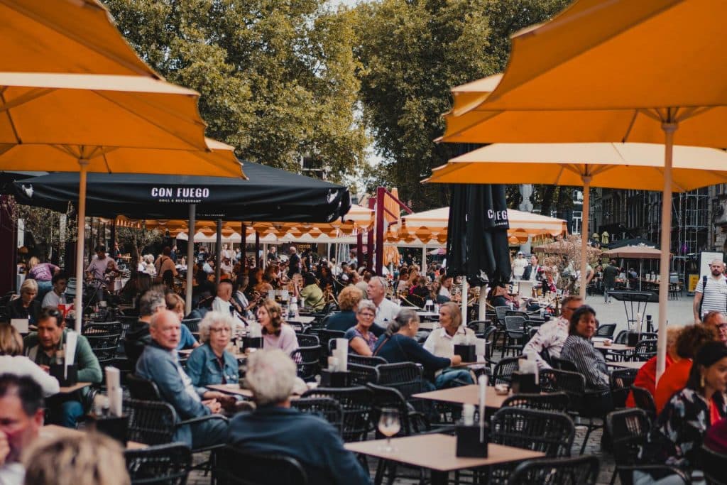 Een terras waar op een drukke dag veel mensen aan tafeltjes onder grote gele parasols zitten en genieten van het goede leven. De levendige sfeer weerspiegelt de geest van Bruisend Breda, terwijl op de achtergrond bomen en gebouwen te zien zijn.