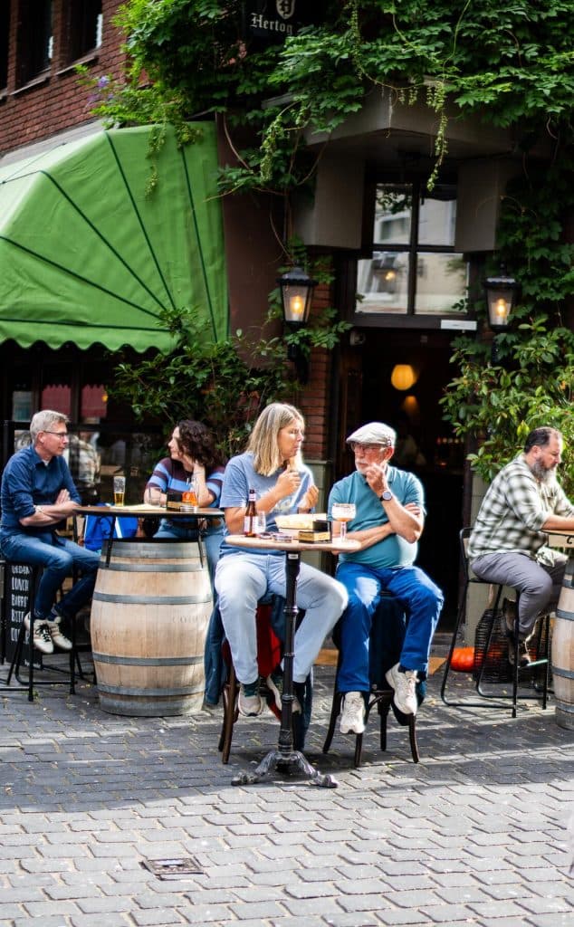 Mensen zitten aan tafeltjes buiten bij een café, drinken en praten – en genieten van het goede leven. Op de foto zijn groen, vaten die als tafels dienen en op de achtergrond een gevel met ramen en lampen te zien, waarmee de levendige sfeer van Bruisend Breda perfect wordt weergegeven.