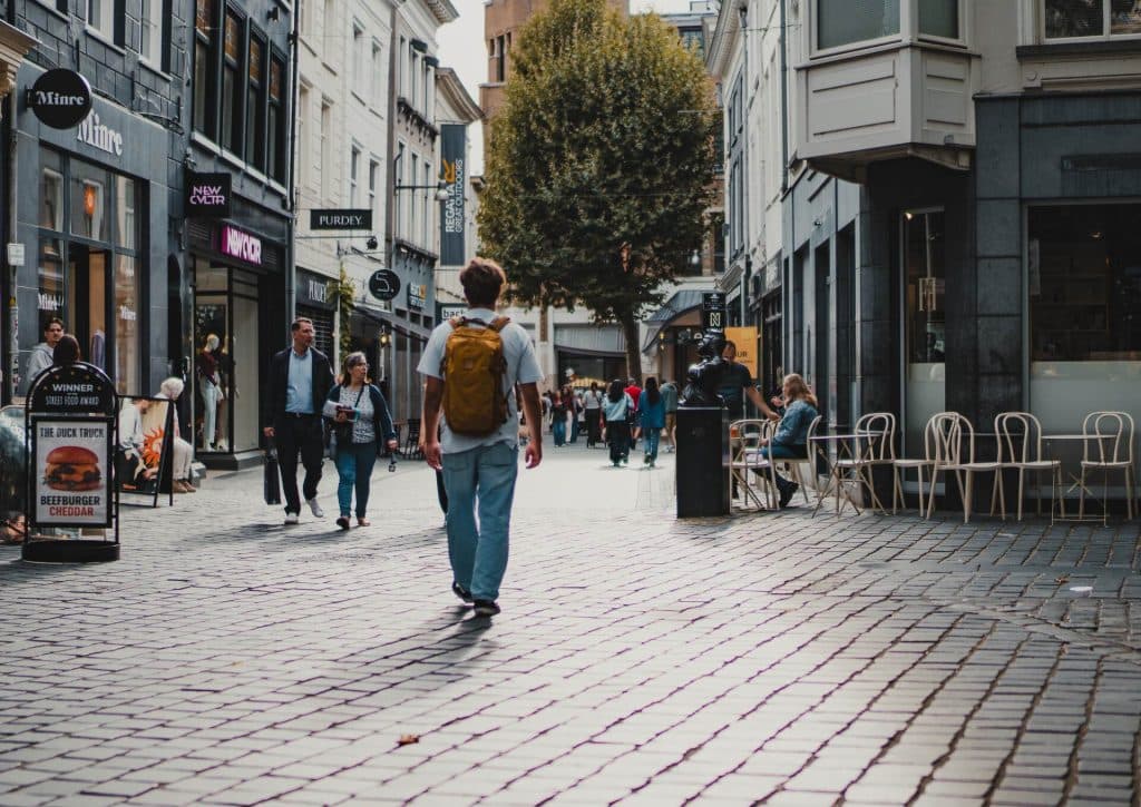 Op een bewolkte dag in Breda loopt iemand met een gele rugzak door een straat met kinderkopjes, waar winkels en terrasjes aan weerszijden staan, en geniet van het goede leven dat de stad te bieden heeft.