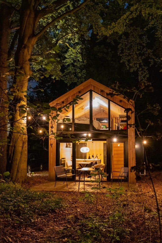 Een klein, houten huisje van twee verdiepingen met grote ramen is ’s avonds verlicht, omringd door bomen, met lichtslingers en een terras aan de voorkant – perfect om onder de sterrenhemel van het goede leven te genieten.