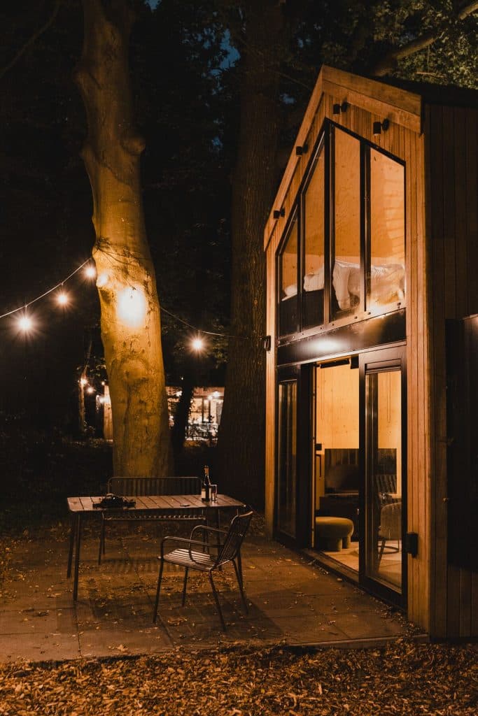 Een moderne houten hut met grote ramen en buitenverlichting. Op het terras staat een tafel met twee stoelen en flessen nonnenwijn, ’s avonds omringd door bomen — een perfecte setting voor het goede leven.
