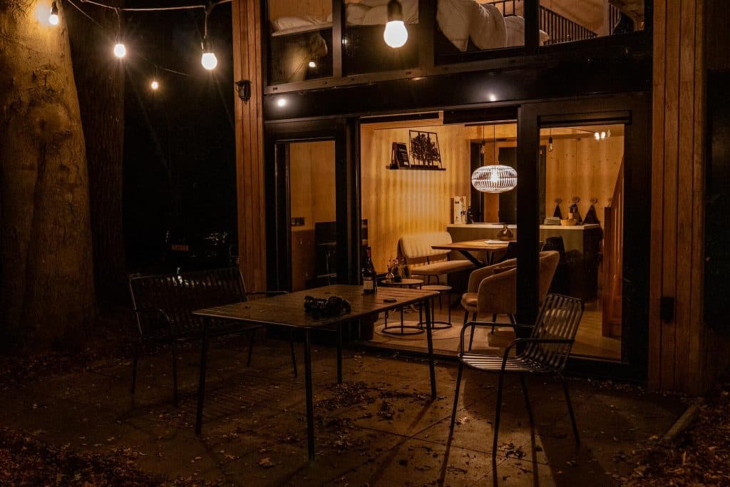 Buitenzicht van een moderne woning bij nacht, met grote glazen deuren, terrasmeubilair, lichtslingers en een warm verlicht interieur dat door de ramen zichtbaar is – een perfect tafereel dat ‘het goede leven’ in Breda perfect weergeeft, aangevuld met een fles nonnenwijn op tafel.