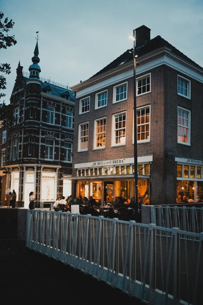 Bij het vallen van de avond zitten mensen aan tafels buiten voor een verlicht restaurant genaamd „De Botanist Breda” in een bakstenen gebouw; ze genieten van het goede leven terwijl ze worden omringd door de levendige sfeer van Bruisend Breda, met een sfeervol historisch gebouw in de buurt en een brug op de voorgrond.