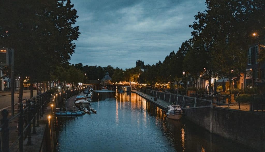 Een gracht in de schemering met aan weerszijden aangemeerde bootjes, bomen en gebouwen langs de oever, en straatlantaarns die in het water weerspiegelen – een perfect tafereel dat ‘het goede leven’ in het bruisende Breda perfect weergeeft.