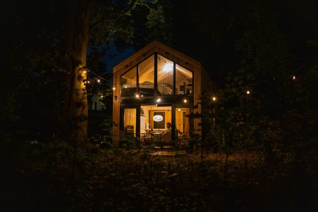 Een moderne hut met grote ramen is ’s avonds verlicht, omringd door bomen net buiten Breda. Binnen zit iemand aan een tafel te genieten van het goede leven, terwijl buiten lichtslingers en de zachte gloed van nonnenwijn een uitnodigende sfeer creëren.