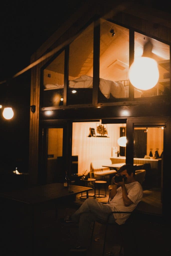 ’s Avonds zit iemand aan een tafel buiten bij een modern huis, verlicht door hanglampen, terwijl er warm licht uit het interieur schijnt. Omringd door het rustige bos bij Breda nipt hij van een glas nonnenwijn en geniet hij van de vredige avond.