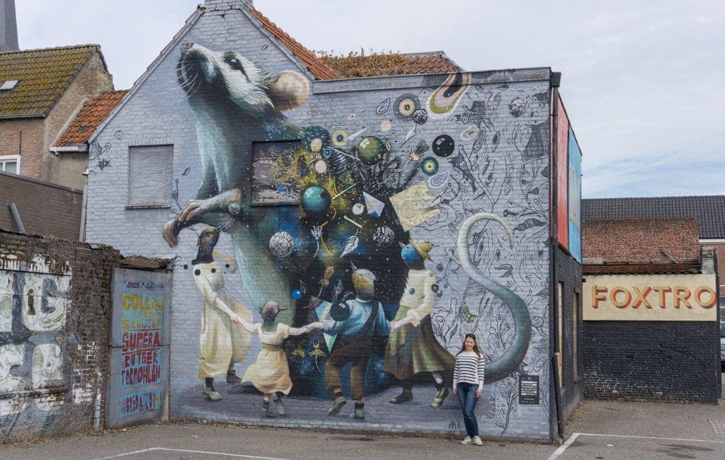 Op een muurschildering aan de gevel van een gebouw zijn een grote witte rat, kinderen in ouderwetse kleding, ronddraaiende planeten en een persoon te zien die ervoor staat te poseren voor de foto – waarmee de sfeer van ‘het goede leven’ wordt weergegeven die Bruisend Breda zo uniek maakt.