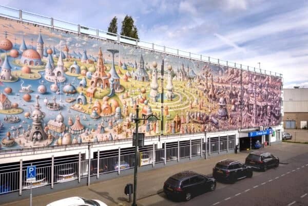 Een grote, kleurrijke muurschildering op de zijkant van een parkeergarage in Breda toont een fantasielandschap met grillige gebouwen en torens onder een gedeeltelijk bewolkte hemel. Het tafereel roept de sfeer van ‘het goede leven’ op en nodigt voorbijgangers uit om zich voor te stellen hoe ze genieten van de geneugten van het leven – misschien zelfs van een glas nonnenwijn – te midden van de betoverende architectuur. Voor de parkeergarage staan auto’s langs de straat geparkeerd.