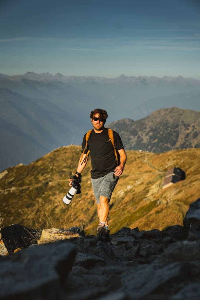 Een man met een zonnebril en een camera wandelt over de rotsen op de Monte Gambarogno, met de Alpen in de verte.