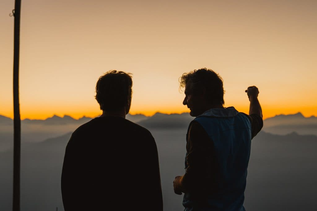 Twee mensen tekenen zich als silhouet af tegen de oranje lucht na zonsondergang, met de bergtoppen van Ticino erachter.