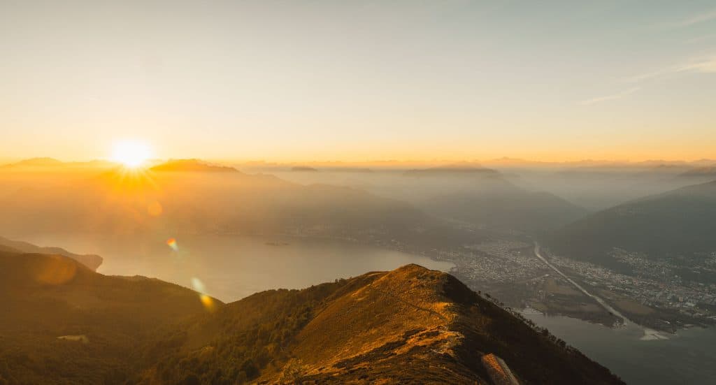 De zon gaat onder boven het Lago Maggiore, gezien vanaf de Monte Gambarogno, met de Maggiadelta en de bergen in de nevel.