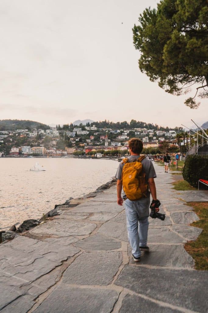 Een man met een gele rugzak en een camera wandelt over de stenen oever van het Lago Maggiore richting Ascona, met de heuvels erachter.