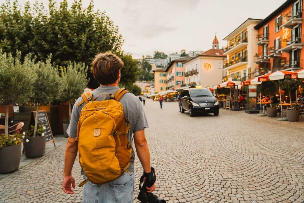 Een man met een gele rugzak loopt over de kasseien van de lungolago in Ascona, langs pastelkleurige gevels en terrassen met parasols.