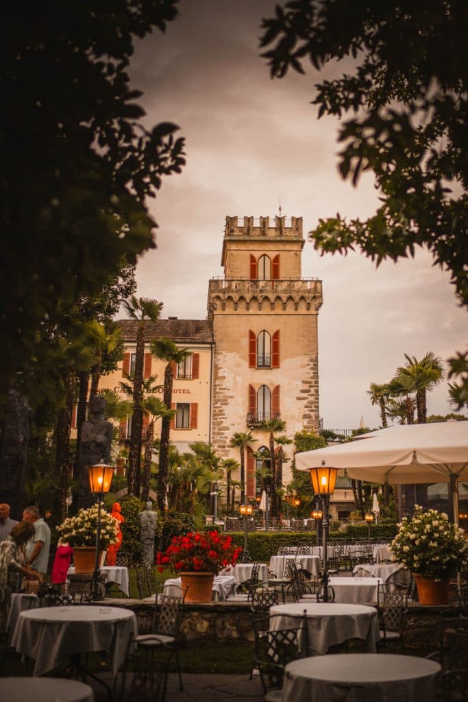 Een oude kasteeltoren tussen palmbomen en bloembakken aan de lungolago van Ascona, met gedekte tafels op het terras ervoor.