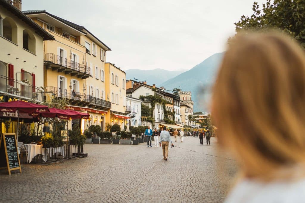 De promenade van Ascona met gele en witte gevels, rode parasols en wandelaars, met de bergen rond het Lago Maggiore op de achtergrond.