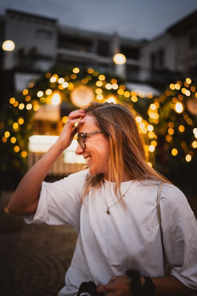 Een lachende vrouw met een bril en een camera staat 's avonds in Ascona voor een boog van warme lichtjes.