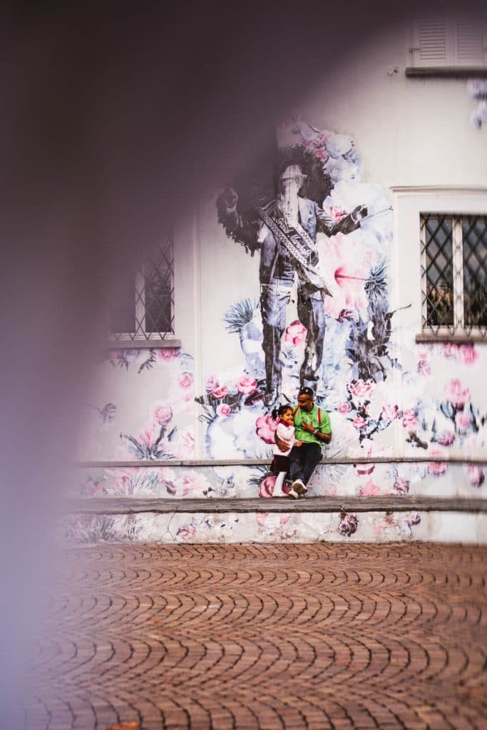 Een grote muurschildering met een figuur en roze bloemen op een witte gevel in Ascona, met twee mensen op een bankje eronder.