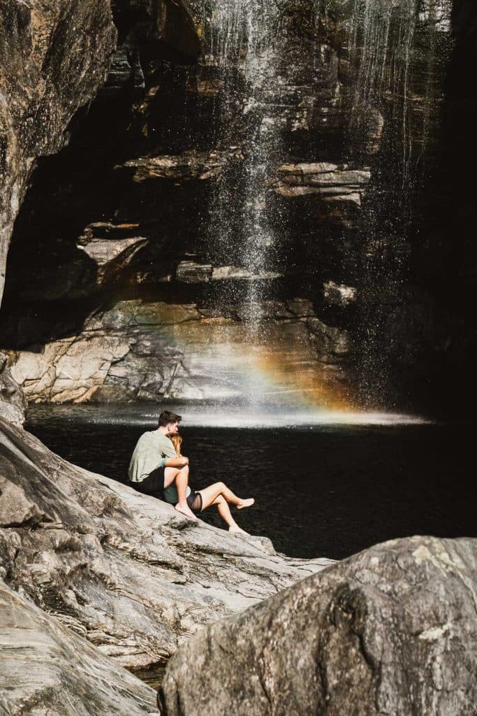 Twee mensen zitten op de rotsen bij de Cascata del Salto in Maggia, waar een regenboog in de nevel van de waterval hangt.