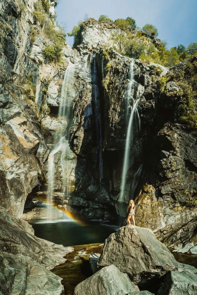 Een smalle waterval stort tussen steile rotsen naar beneden in de Maggiavallei, met een regenboog boven de kom en een persoon op een rotsblok.