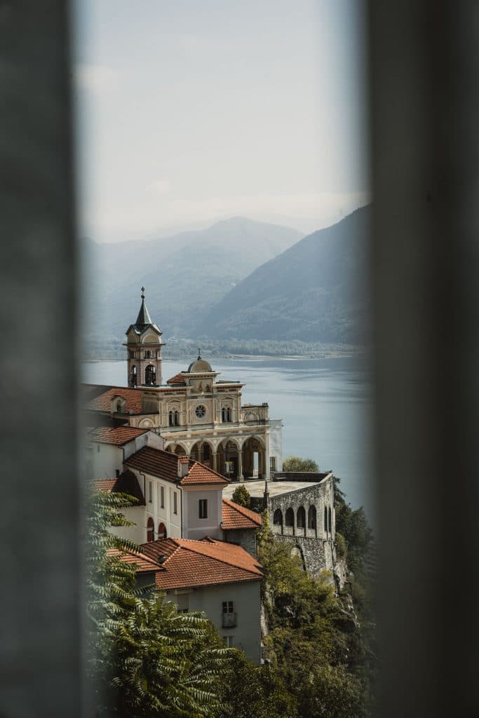 Het heiligdom Madonna del Sasso, met zijn okergele kerk en klokkentoren, tussen twee palen door gezien, met het Lago Maggiore erachter.